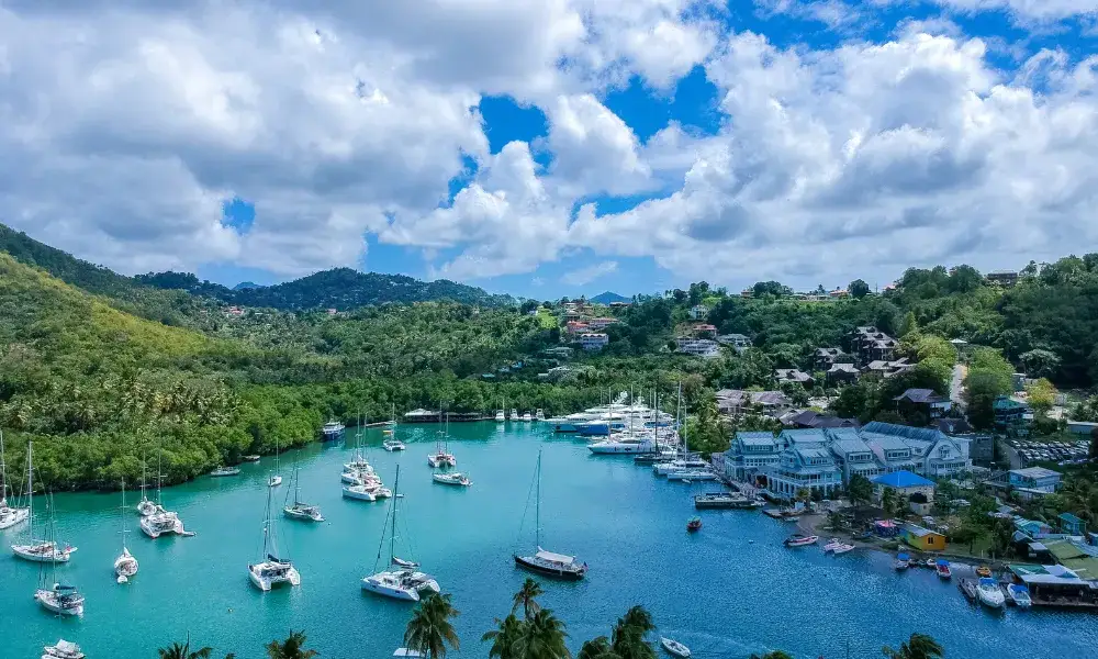 A stunning aerial view of a picturesque bay in Saint Lucia, featuring turquoise waters, anchored sailboats, lush green hills, and a charming marina with waterfront buildings under a bright blue sky with fluffy clouds