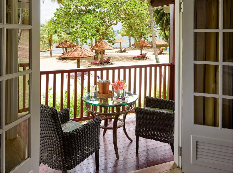 A private cottage balcony at East Winds, St. Lucia, featuring a cozy seating area, a table set with chilled champagne and flowers, overlooking a serene beachfront with thatched cabanas