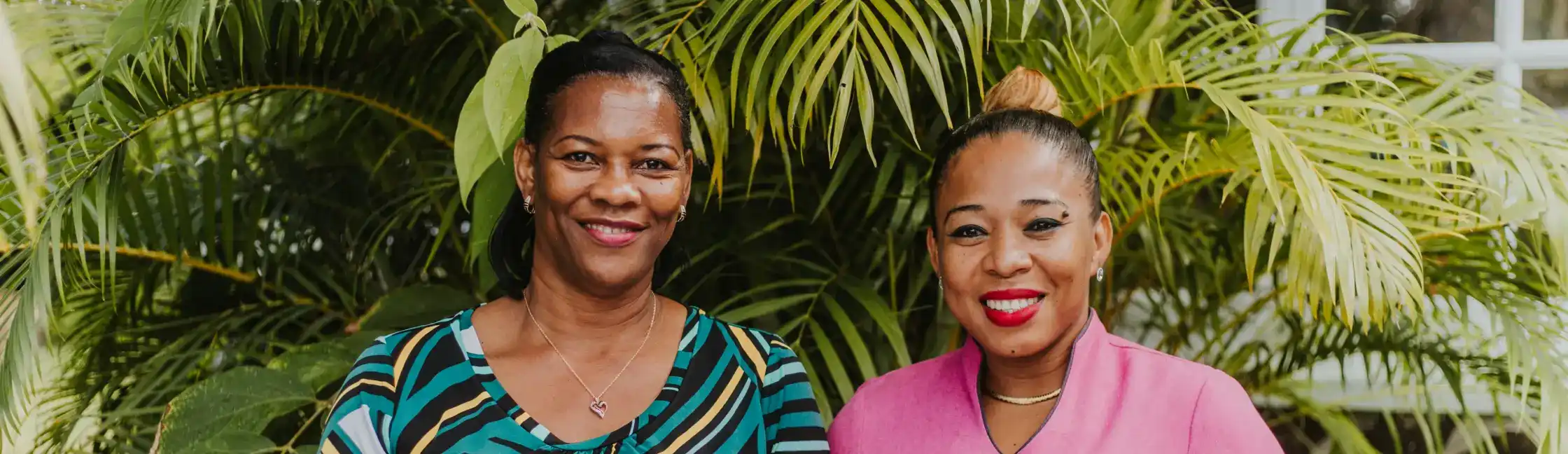 Two women smiling in front of lush green tropical foliage.