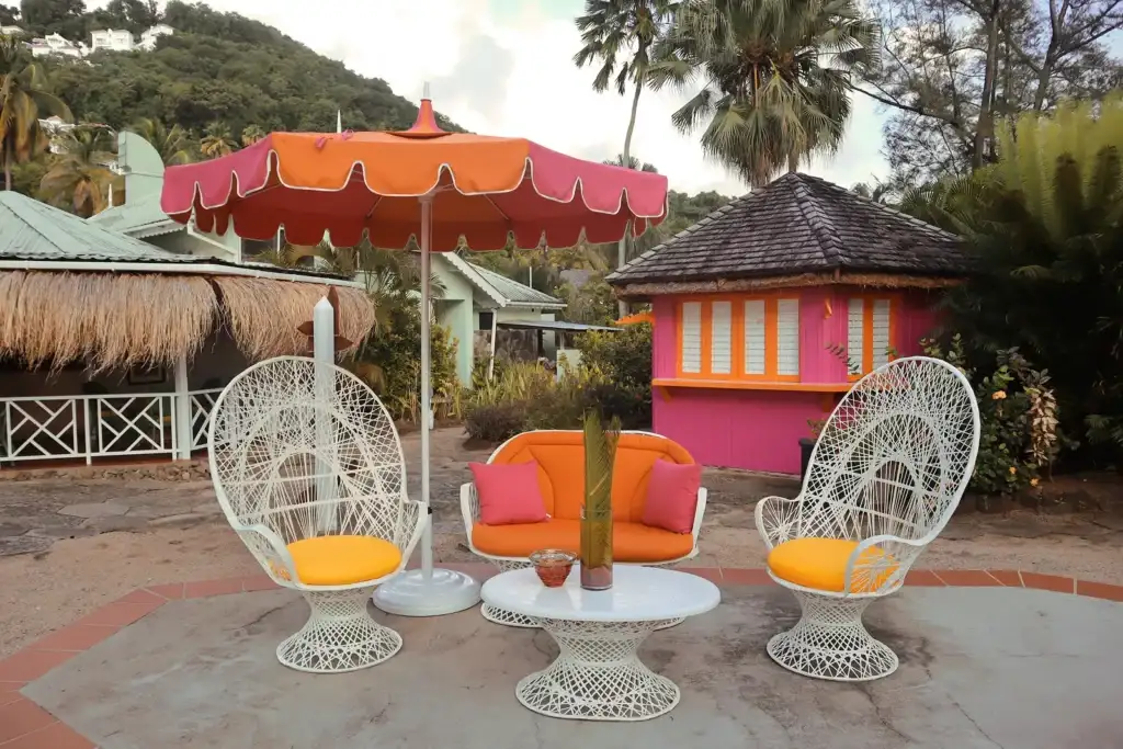 A vibrant outdoor seating area at East Winds Resort, St. Lucia, featuring elegant white wicker chairs, a tropical umbrella, and colorful Caribbean-style cottages in a lush, serene setting.