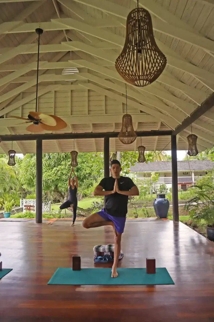 A serene open-air yoga pavilion at East Winds Resort, St. Lucia, surrounded by lush greenery. Guests engage in a peaceful yoga session, embracing wellness and tranquility in a tropical setting.