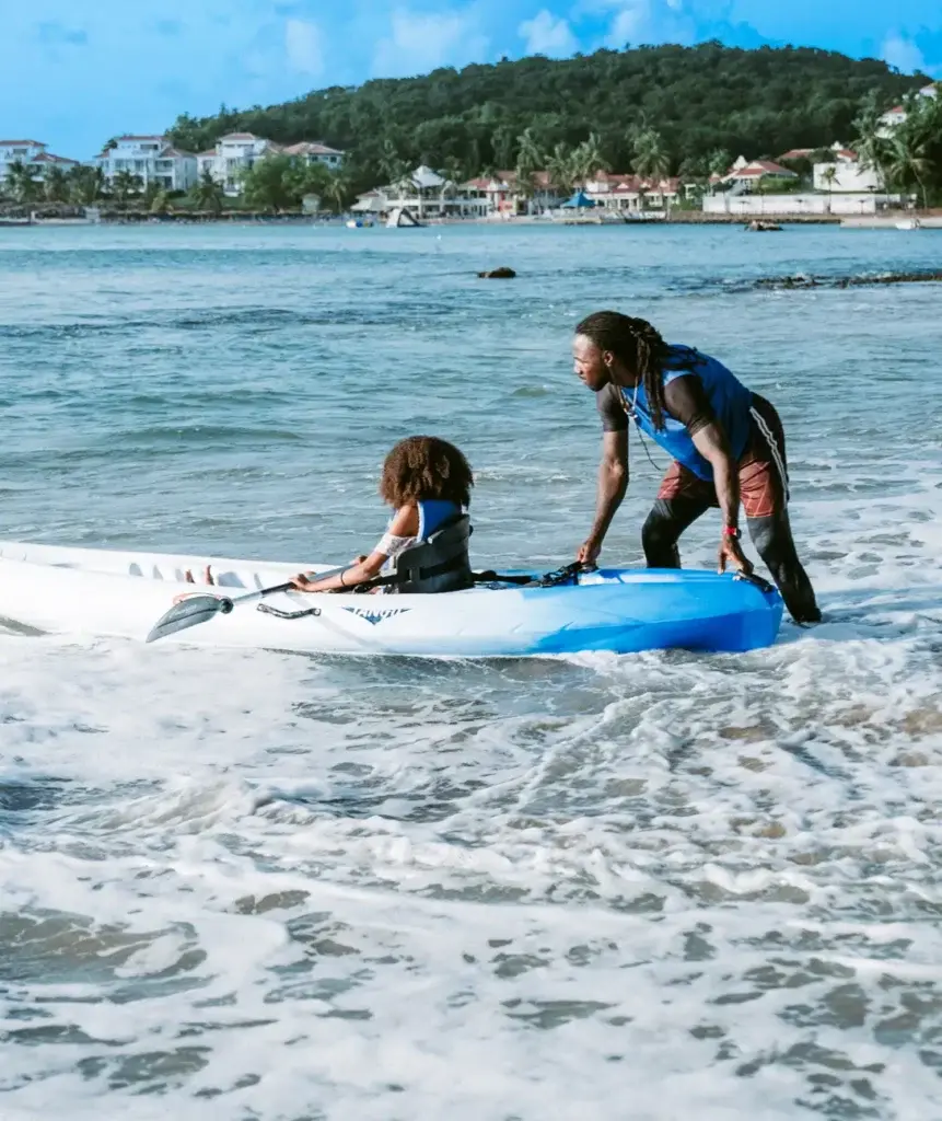Child kayaking with adult help on a beach with clear blue water and lush greenery.